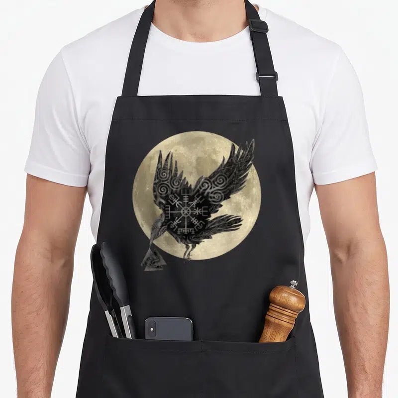 Tablier de cuisine noir avec corbeau stylisé, motifs runiques et pleine lune en arrière-plan beige.
