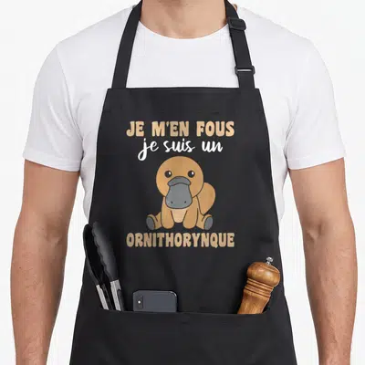 Tablier de cuisine noir avec un dessin d'ornithorynque marron et le texte Je m'en fous je suis un ornithorynque en beige et blanc.