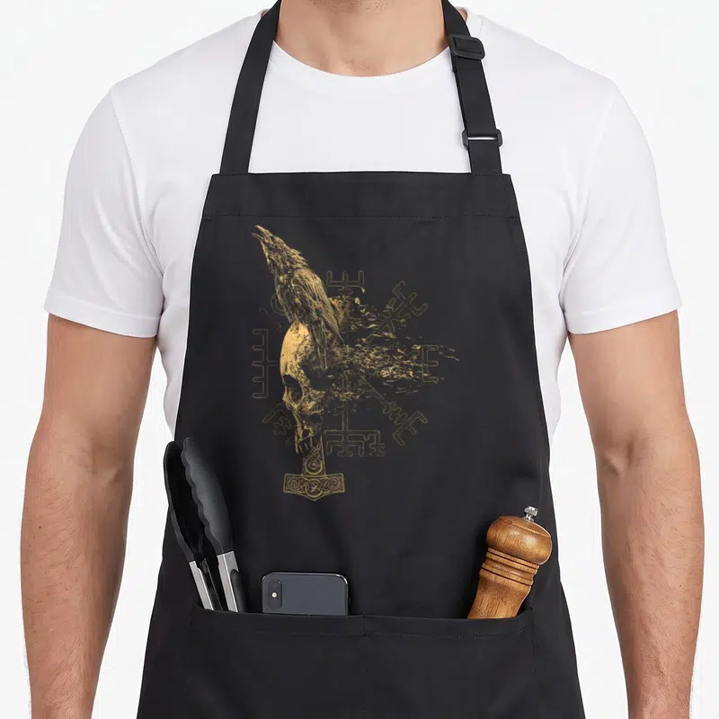Tablier de cuisine noir avec illustration dorée d'un corbeau, un crâne, le marteau de Thor et le Vegvisir.