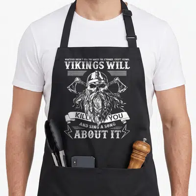Tablier de cuisine noir avec illustration blanche d'un viking crâne à haches et texte humoristique sur fond uni.