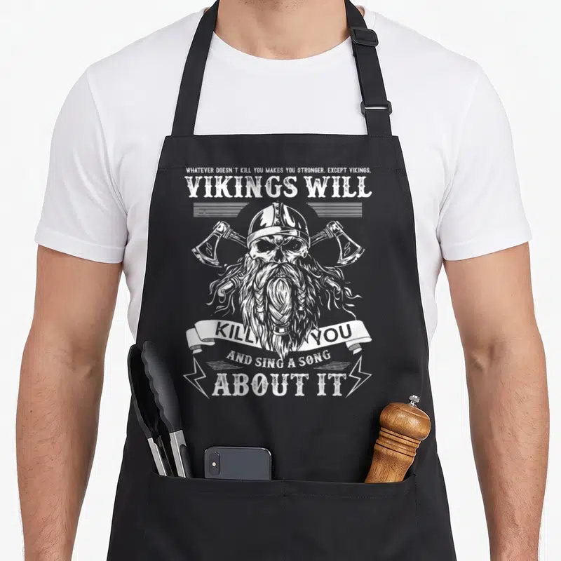 Tablier de cuisine noir avec illustration blanche d'un viking crâne à haches et texte humoristique sur fond uni.