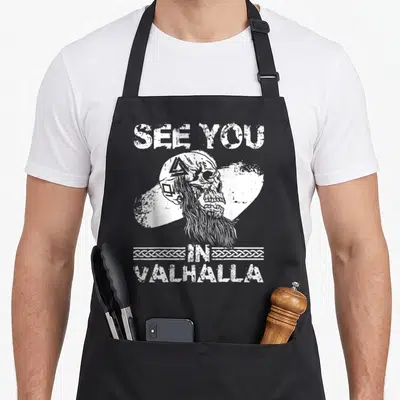 Tablier de cuisine noir avec un crâne viking barbu, le texte See You In Valhalla et des motifs de tresses scandinaves en blanc.