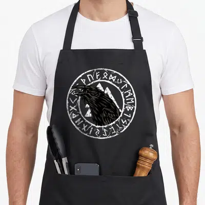 Tablier de cuisine noir avec tête de corbeau et runes celtiques en cercle blanc.