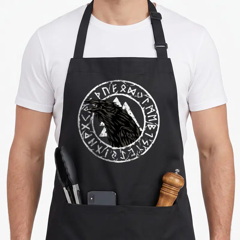 Tablier de cuisine noir avec tête de corbeau et runes celtiques en cercle blanc.