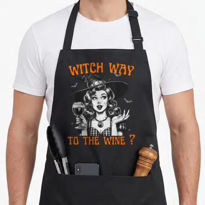 Tablier de cuisine noir avec illustration sorcière pin-up tenant un verre de vin et texte orange Witch Way To The Wine.