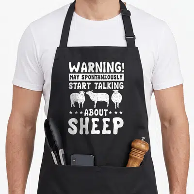 Tablier de cuisine noir avec l'inscription Warning May Spontaneously Start Talking About Sheep et trois moutons dessinés en blanc.