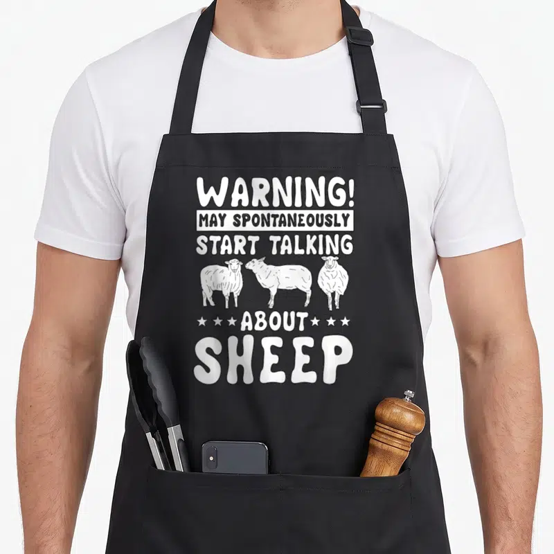 Tablier de cuisine noir avec l'inscription Warning May Spontaneously Start Talking About Sheep et trois moutons dessinés en blanc.