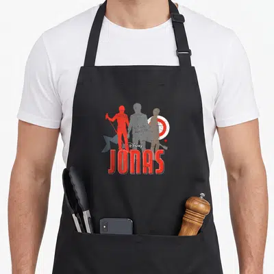 Tablier de cuisine noir avec les silhouettes rouges et grises du groupe JONAS et le logo Disney au centre.