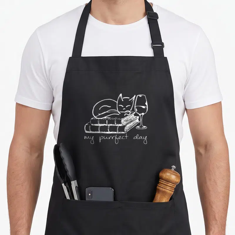 Tablier de cuisine noir avec illustration blanche d'un chat sur des livres et un verre de vin, texte my purrfect day