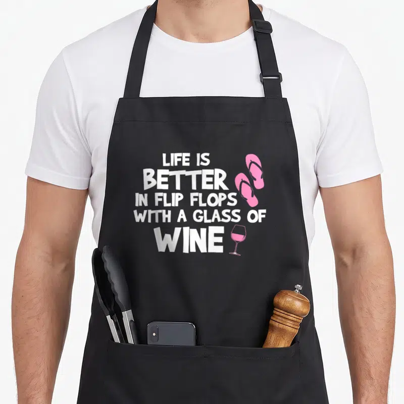 Tablier de cuisine noir avec texte blanc Life is Better in Flip Flops with a Glass of Wine et illustrations roses de tongs et verre de vin.