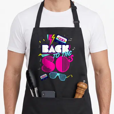 Tablier de cuisine noir avec inscription Back to the 80's, cassettes, lunettes shutter et motifs géométriques roses et bleus.