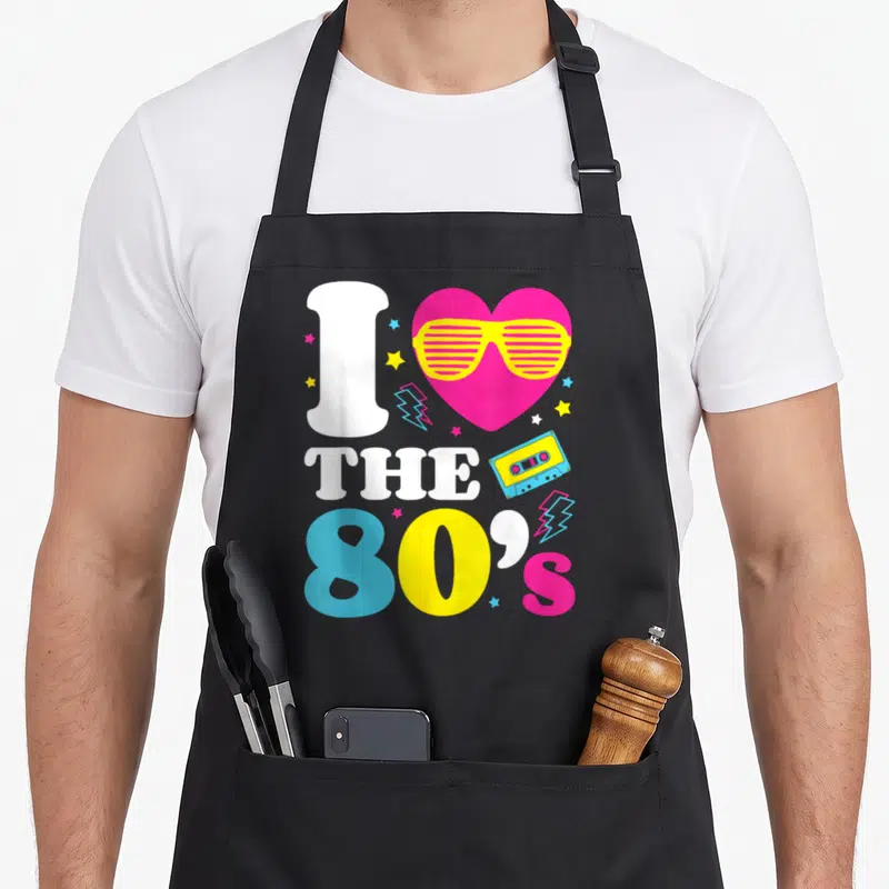 Tablier de cuisine noir avec un grand cœur rose portant des lunettes jaunes et le texte I Love The 80's en lettres colorées.