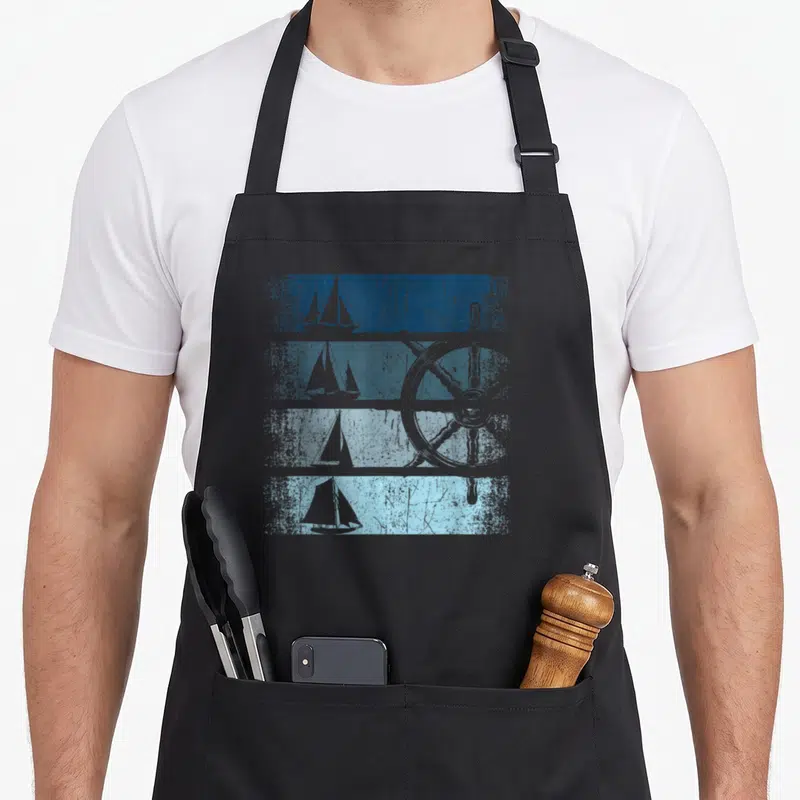 Tablier de cuisine noir avec illustration rétro de voiliers et roue de bateau sur fond bleu texturé