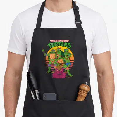 Tablier de cuisine noir avec illustration des tortues ninja et du maître Splinter sur fond de soleil orange rayé.