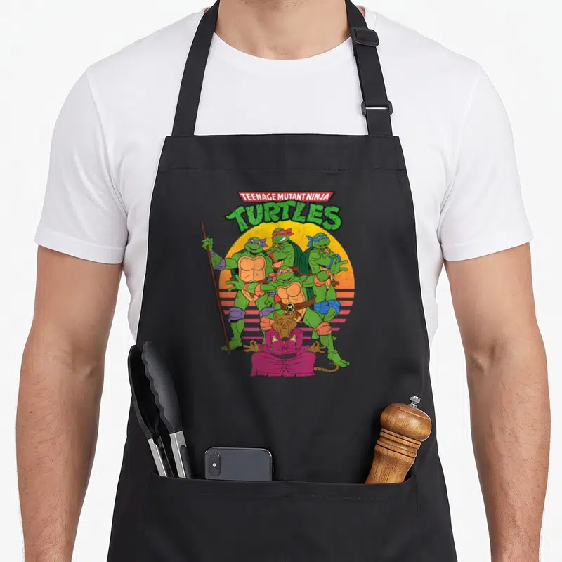 Tablier de cuisine noir avec illustration des tortues ninja et du maître Splinter sur fond de soleil orange rayé.
