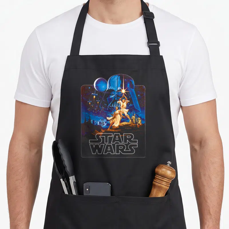 Tablier de cuisine noir avec illustration rétro Star Wars montrant Darth Vader et les personnages principaux sur fond d'espace bleu.