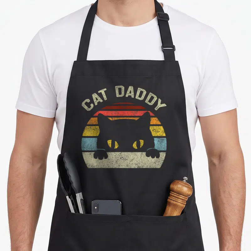 Tablier de cuisine noir avec illustration rétro d'un chat gris et du texte Cat Daddy sur fond de soleil multicolore.