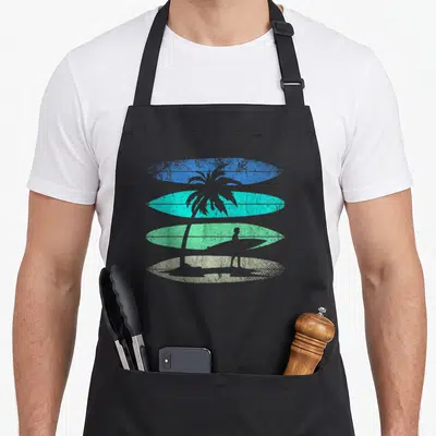 Tablier de cuisine noir avec illustration rétro de planches de surf et silhouette de surfeur sous un palmier.