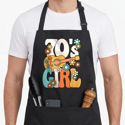 Tablier de cuisine noir avec l'inscription 70's Girl, une guitare acoustique orange et des fleurs multicolores dans un style hippie rétro.