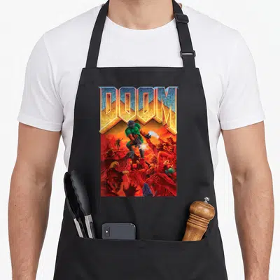 Tablier de cuisine noir avec illustration colorée d'un soldat vert combattant des démons rouges sous un logo DOOM géométrique.