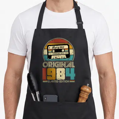 Tablier de cuisine noir avec illustration de cassette audio rétro et le texte Original 1984 Limited Edition en couleurs pastel.