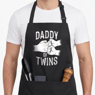 Tablier de cuisine noir avec inscription Daddy of Twins et illustration de deux mains effectuant un poing serré.