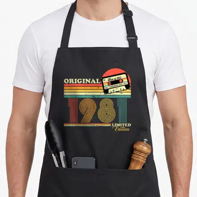 Tablier de cuisine noir avec illustration cassette audio rétro et les chiffres 1981 en lettres colorées.