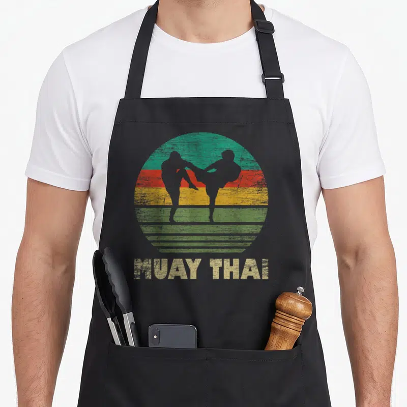 Tablier de cuisine noir avec illustration rétro de deux silhouettes en Muay Thai sur un soleil aux bandes vertes, rouges et jaunes.