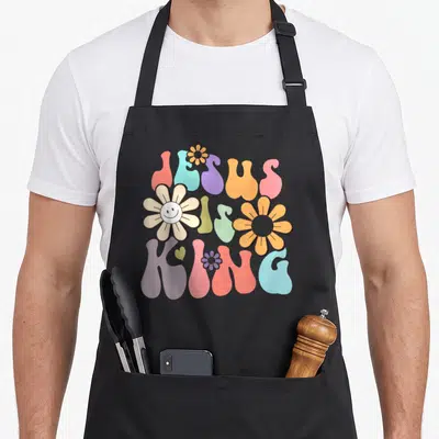 Tablier de cuisine noir avec inscription Jésus est Roi en lettres pastels et fleurs stylisées.