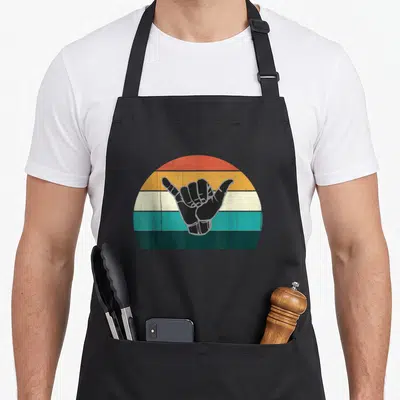 Tablier de cuisine noir avec une main Shaka noire sur un demi-cercle aux bandes colorées orange, jaune, blanc, turquoise et vert foncé.