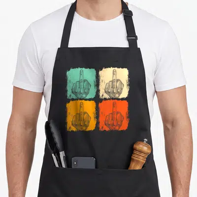 Tablier de cuisine noir avec quatre illustrations de mains levant le majeur sur fonds colorés et texte humoristique.