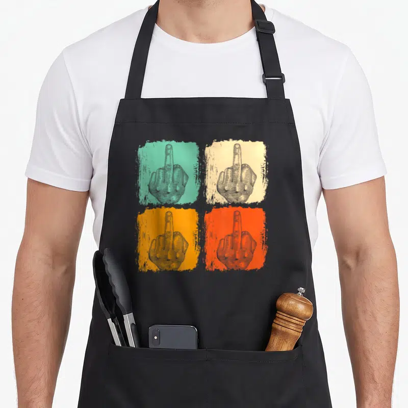 Tablier de cuisine noir avec quatre illustrations de mains levant le majeur sur fonds colorés et texte humoristique.