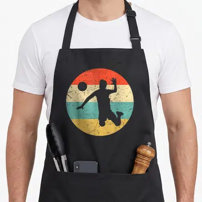 Tablier de cuisine noir avec motif circulaire retro représentant un joueur de volley-ball en silhouette sur bandes colorées délavées.