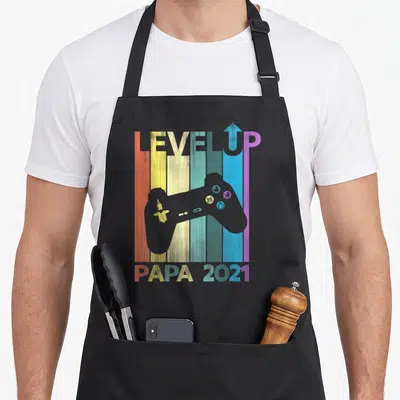 Tablier de cuisine noir avec manette de jeu vidéo colorée et texte Level Up Papa 2021 en style rétro.