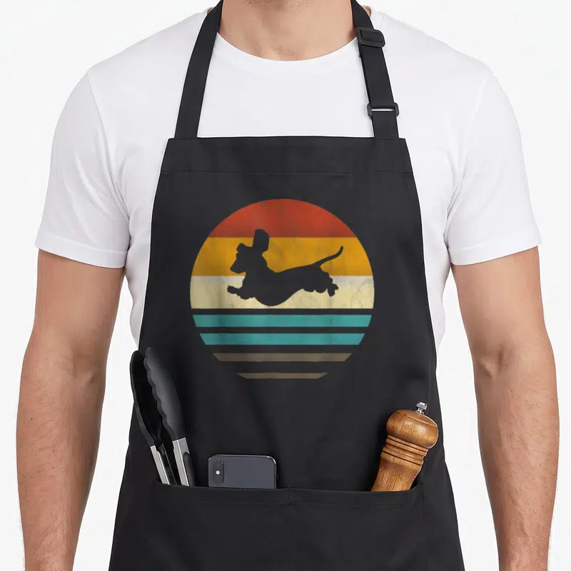 Tablier de cuisine noir avec silhouette de teckel gris sur fond de soleil rayé orange jaune turquoise et beige style rétro.