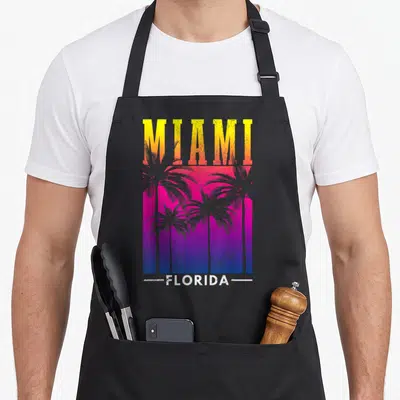 Tablier de cuisine noir avec inscription MIAMI FLORIDA et silhouettes de palmiers sur fond dégradé rose violet bleu.