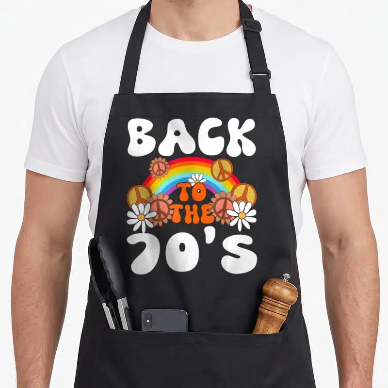 Tablier de cuisine noir avec arc en ciel, symboles de la paix et texte Back to the 70's imprimé en couleurs vives.
