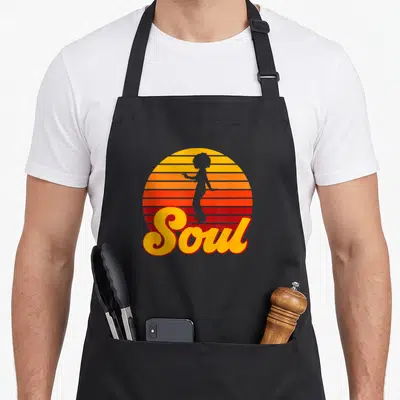 Tablier de cuisine noir avec une silhouette afro sur un soleil strié orange et rouge, le mot Soul en lettres jaunes ondulées.