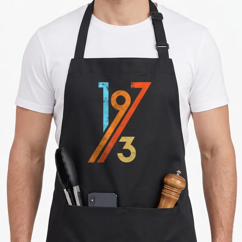 Tablier de cuisine noir avec le motif graphique 1973 aux couleurs bleu orange rouge et jaune sur fond uni.