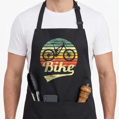 Tablier de cuisine noir avec illustration rétro d'un vélo tout-terrain devant un soleil rayé multicolore et le mot Bike en typographie stylisée.