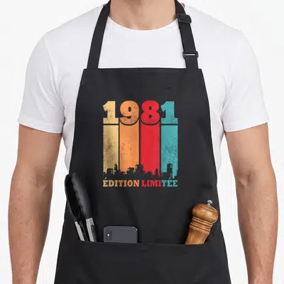 Tablier de cuisine noir avec l'année 1981 en lettres colorées et une silhouette urbaine en bas du design imprimé.