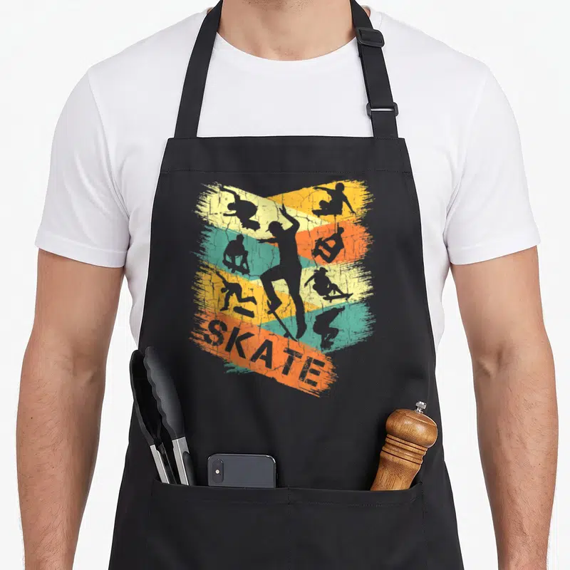 Tablier de cuisine noir avec illustration rétro de skateboarders et le mot SKATE sur bandes colorées.