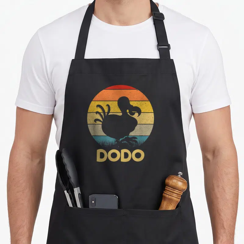 Tablier de cuisine noir avec silhouette de dodo devant un soleil couchant coloré et le texte DODO en bas.