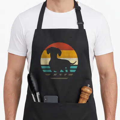 Tablier de cuisine noir avec une silhouette de teckel grise sur un soleil rétro multicolore aux bandes horizontales orange, jaune et turquoise.