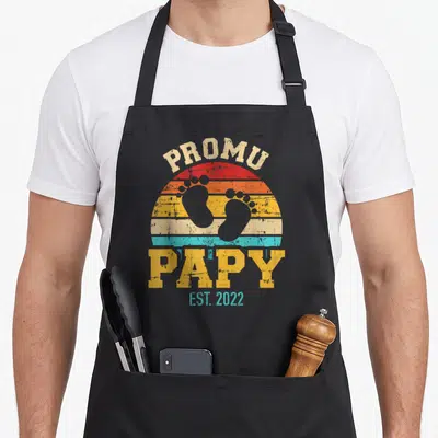 Tablier de cuisine noir avec motif vintage Promu papy Est 2022 et empreintes de pieds de bébé sur fond de soleil coloré.
