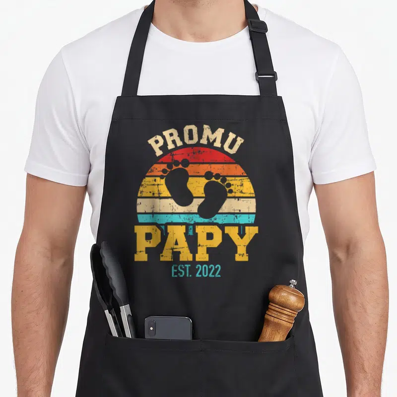 Tablier de cuisine noir avec motif vintage Promu papy Est 2022 et empreintes de pieds de bébé sur fond de soleil coloré.
