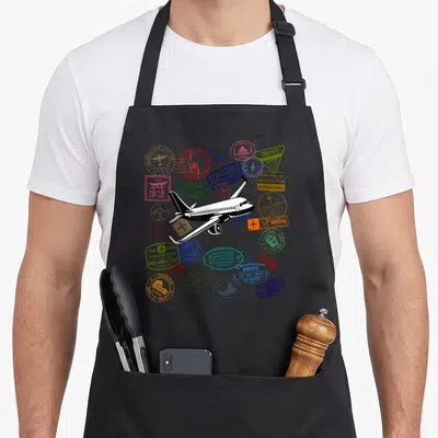 Tablier de cuisine noir avec avion blanc et multiples tampons de voyage colorés sur fond uni.
