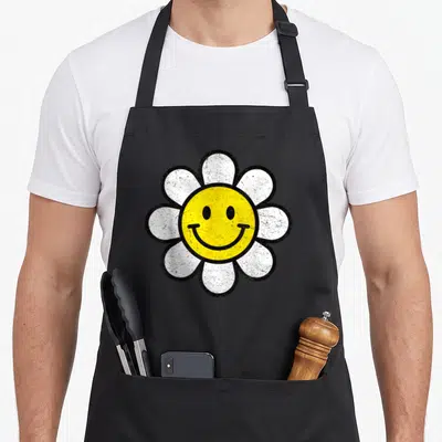 Tablier de cuisine noir avec illustration d'une fleur marguerite blanche au centre jaune portant un visage souriant rétro.