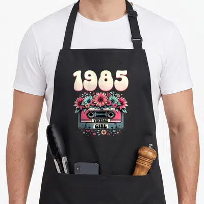 Tablier de cuisine noir avec une cassette rose, des fleurs et l'inscription 1985 Vintage Girl