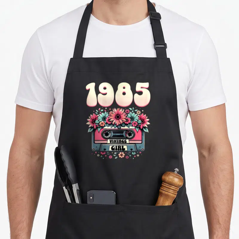 Tablier de cuisine noir avec une cassette rose, des fleurs et l'inscription 1985 Vintage Girl